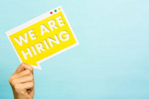 Hand holding on blue background to the left side an advertisement yellow with the message: we are hiring. To the right side is empty for posting a message or copy space.