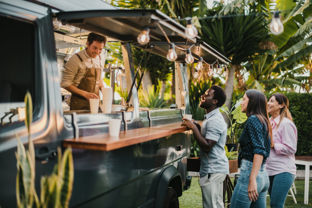 Friends standing in queue and ordering takeaway dishes from man in apron working in food truck in summer in park